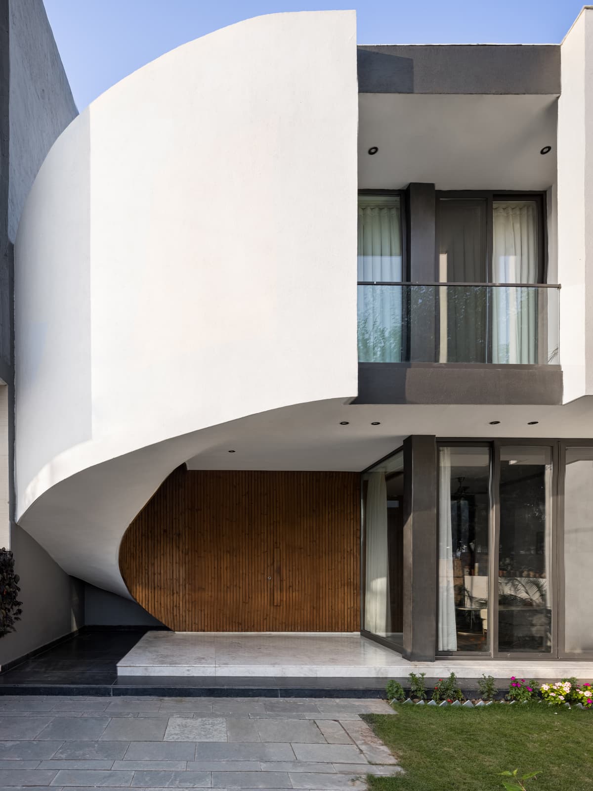 Modern facade with sweeping cantilever curve, vertical timber cladding, recessed lighting, glass balcony railing, and landscaped forecourt emphasizing fluid contemporary form.