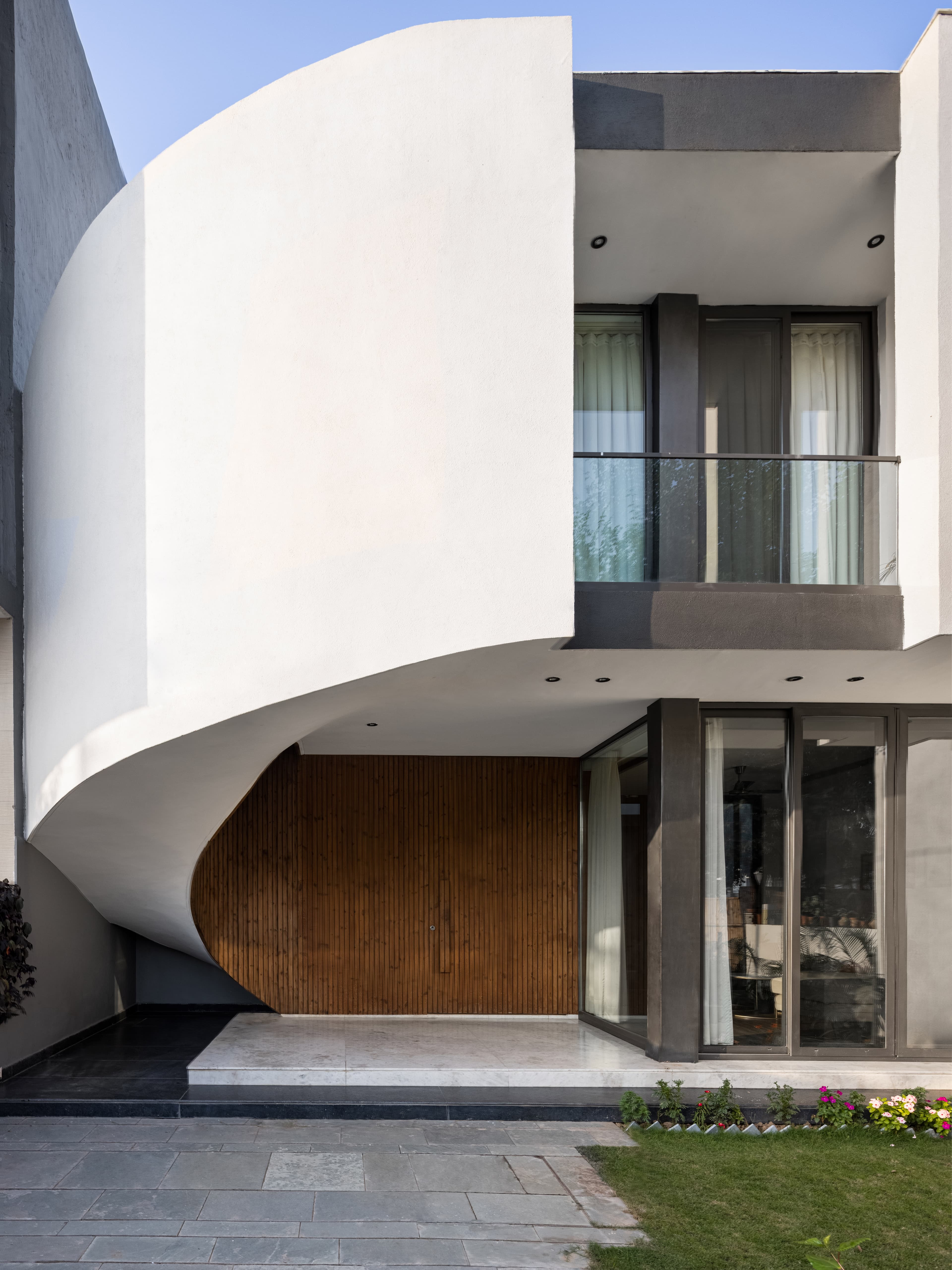 Modern facade with sweeping cantilever curve, vertical timber cladding, recessed lighting, glass balcony railing, and landscaped forecourt emphasizing fluid contemporary form.