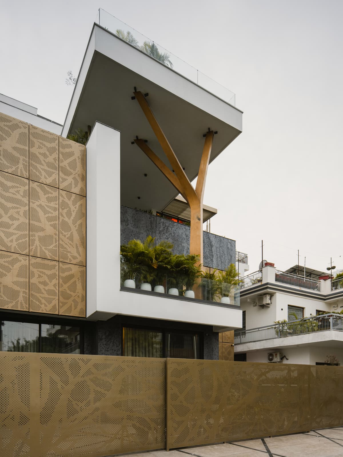 The building showcases a striking Y-shaped column, extended canopy, perforated metal panels, glass railings, and lush planter-lined balcony.