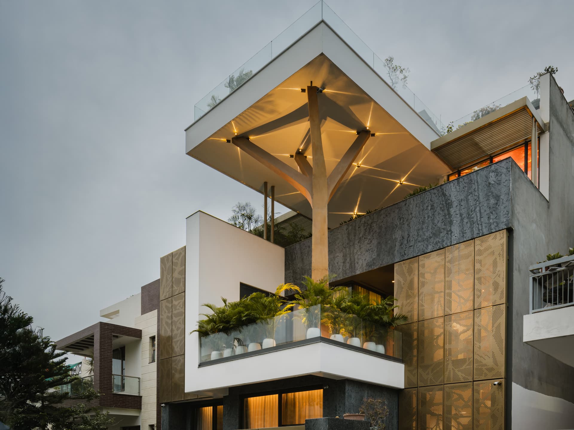 An illuminated canopy supported by a Y-shaped column crowns this home, featuring perforated metal panels, textured stone, and glass railings.