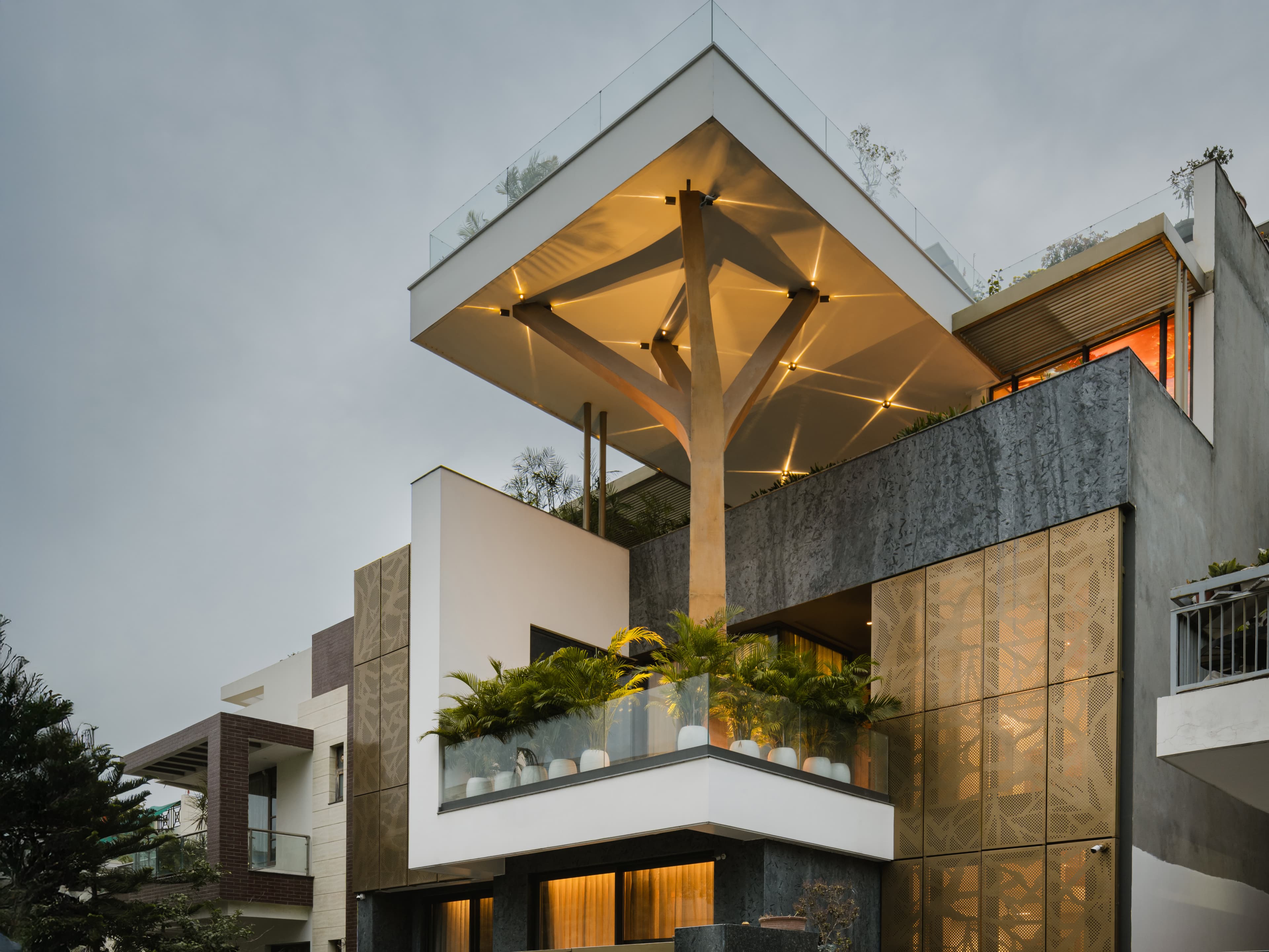An illuminated canopy supported by a Y-shaped column crowns this home, featuring perforated metal panels, textured stone, and glass railings.