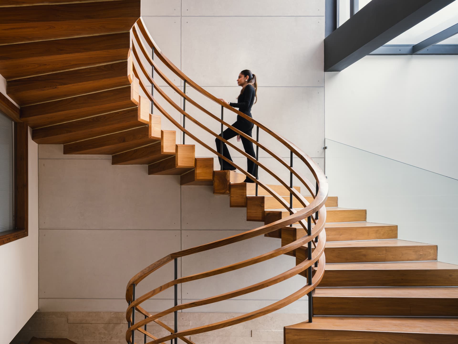 A sleek wooden staircase with curved railings, cantilevered treads, clean concrete walls, and natural light streaming through large skylight windows.