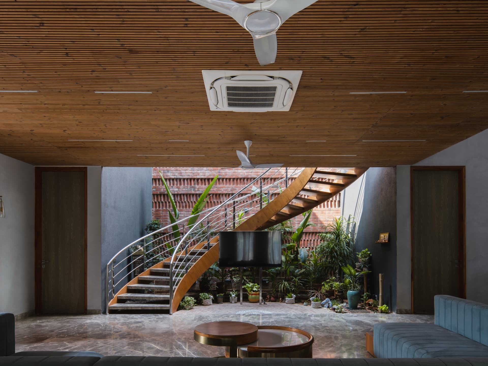Curved wooden staircase, exposed brick jaali, and indoor courtyard blend biophilic design with warm textures and open-plan spatial fluidity.
