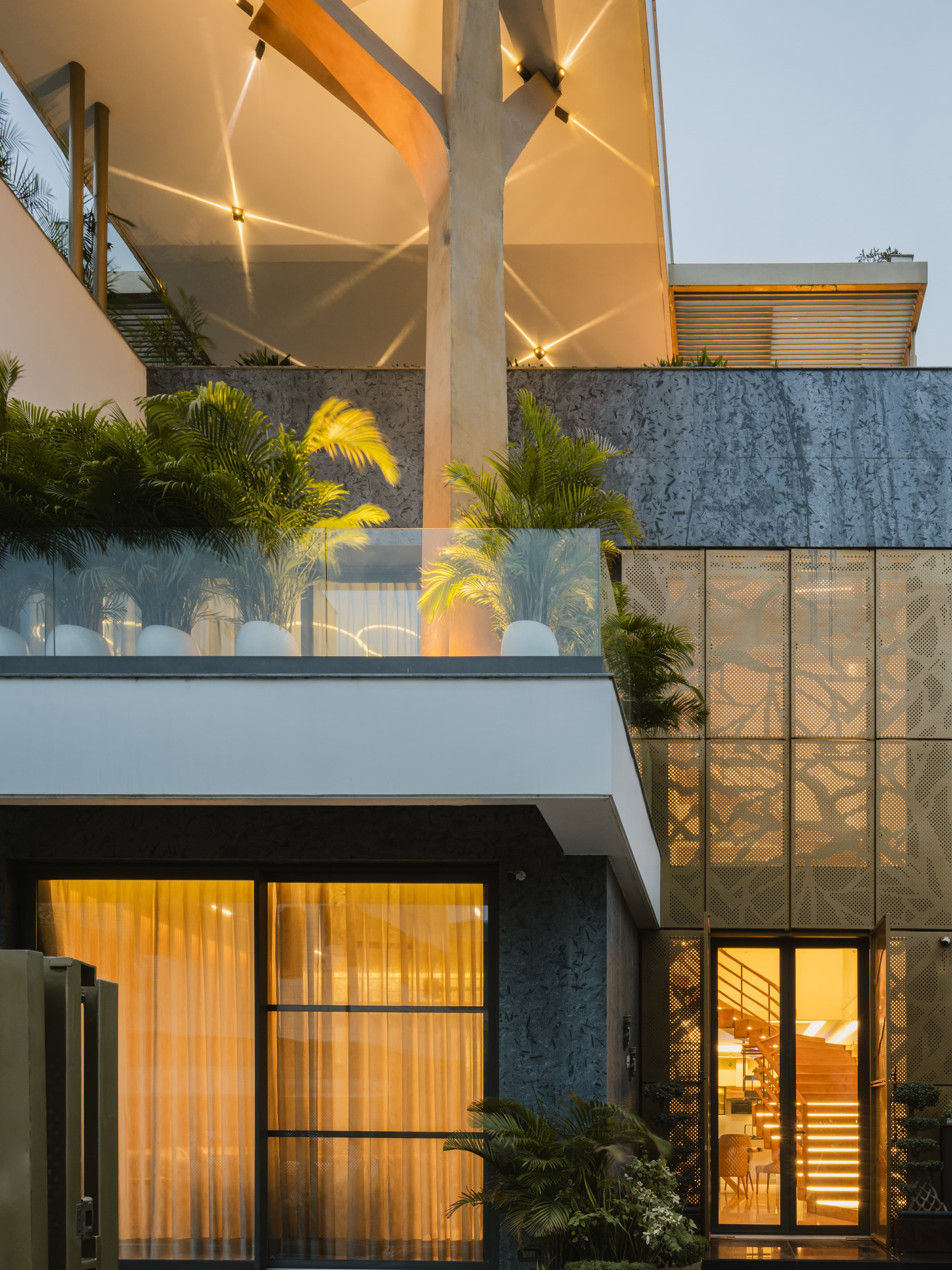 Backlit Y-column supports an illuminated canopy above a lush balcony, with perforated metal cladding, large windows, and a visible staircase.