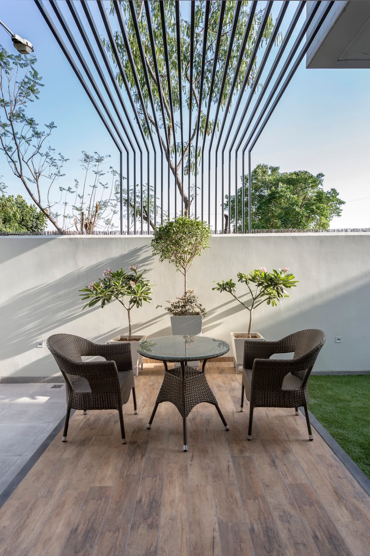 Cozy outdoor patio with wicker furniture, potted greenery, wooden flooring, and striking vertical trellis framing sky views in minimalist elegance.