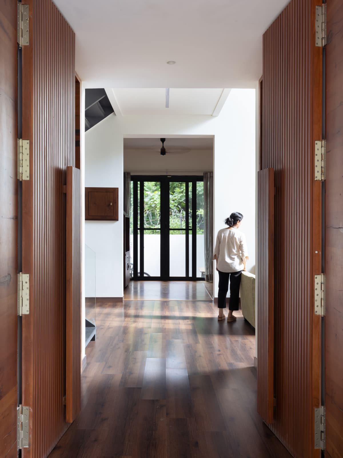 Long corridor with wooden flooring and fluted wooden panel doors; natural lighting leads to a bright living space with large fenestration.