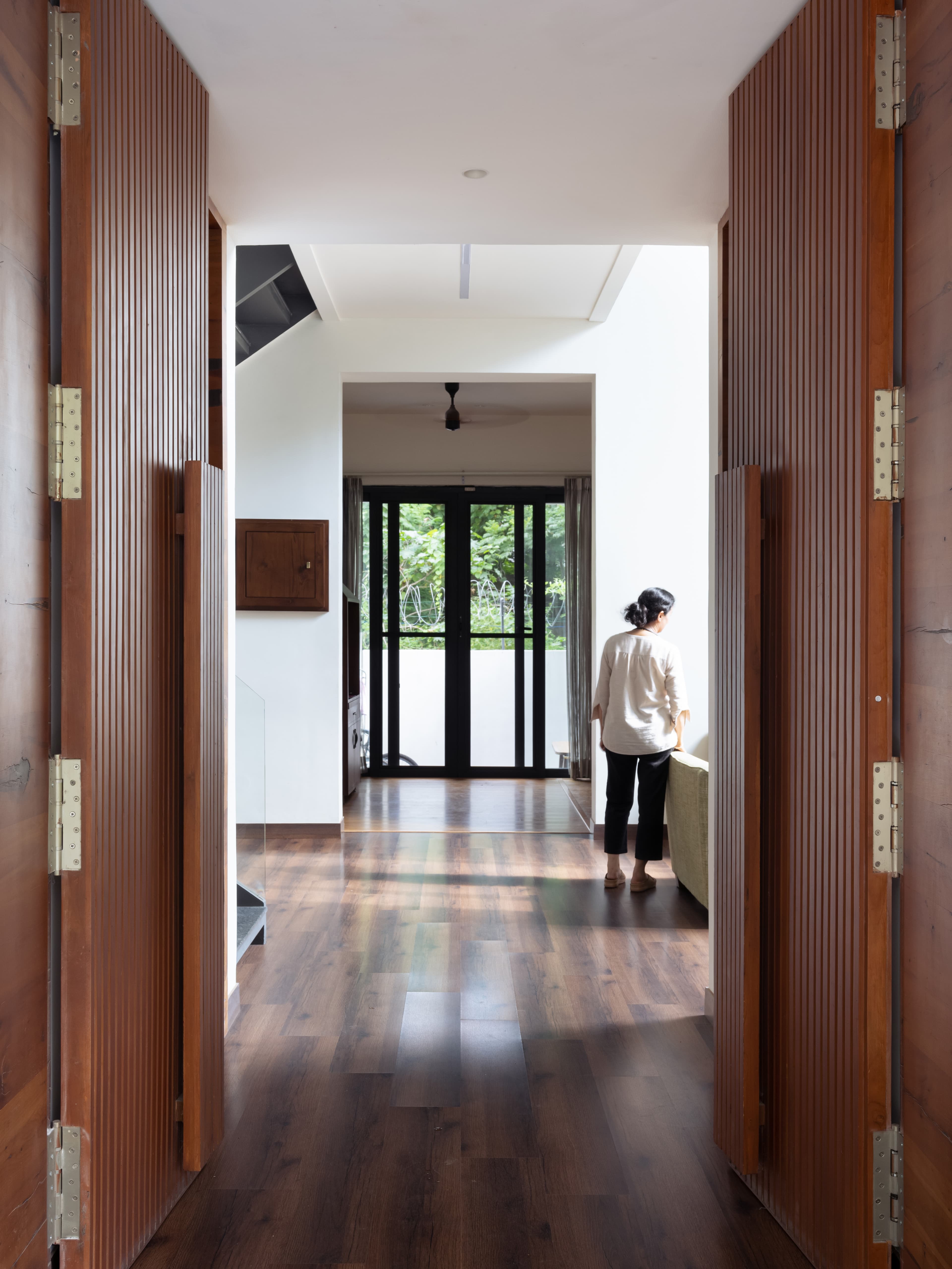Long corridor with wooden flooring and fluted wooden panel doors; natural lighting leads to a bright living space with large fenestration.