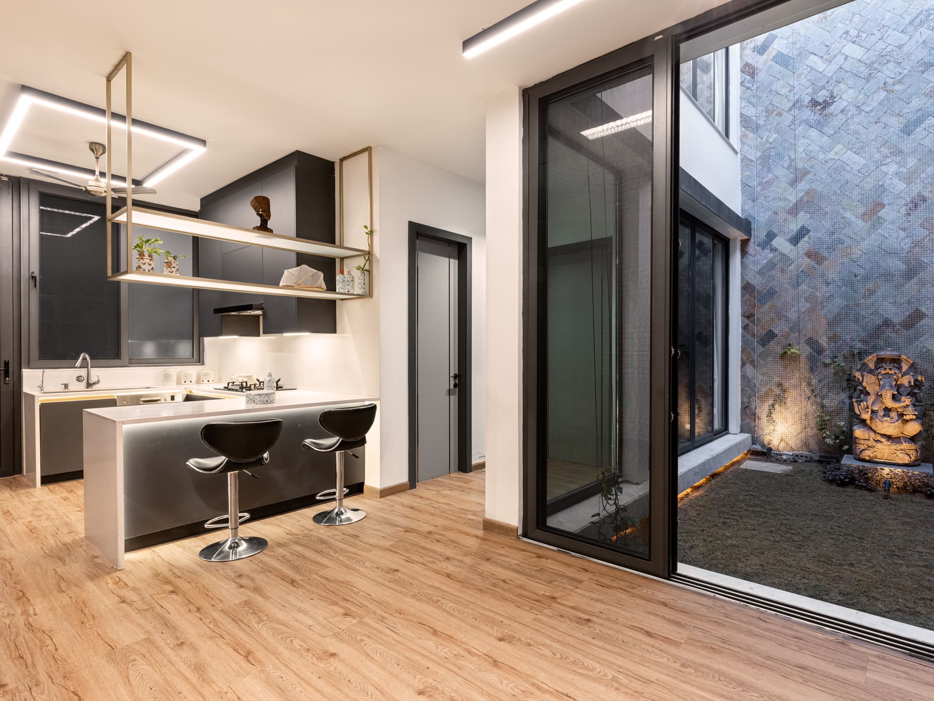Open-plan kitchen with sleek cabinetry, suspended shelving, and large glazing connecting to stone-clad courtyard featuring illuminated sculpture and textured walls.