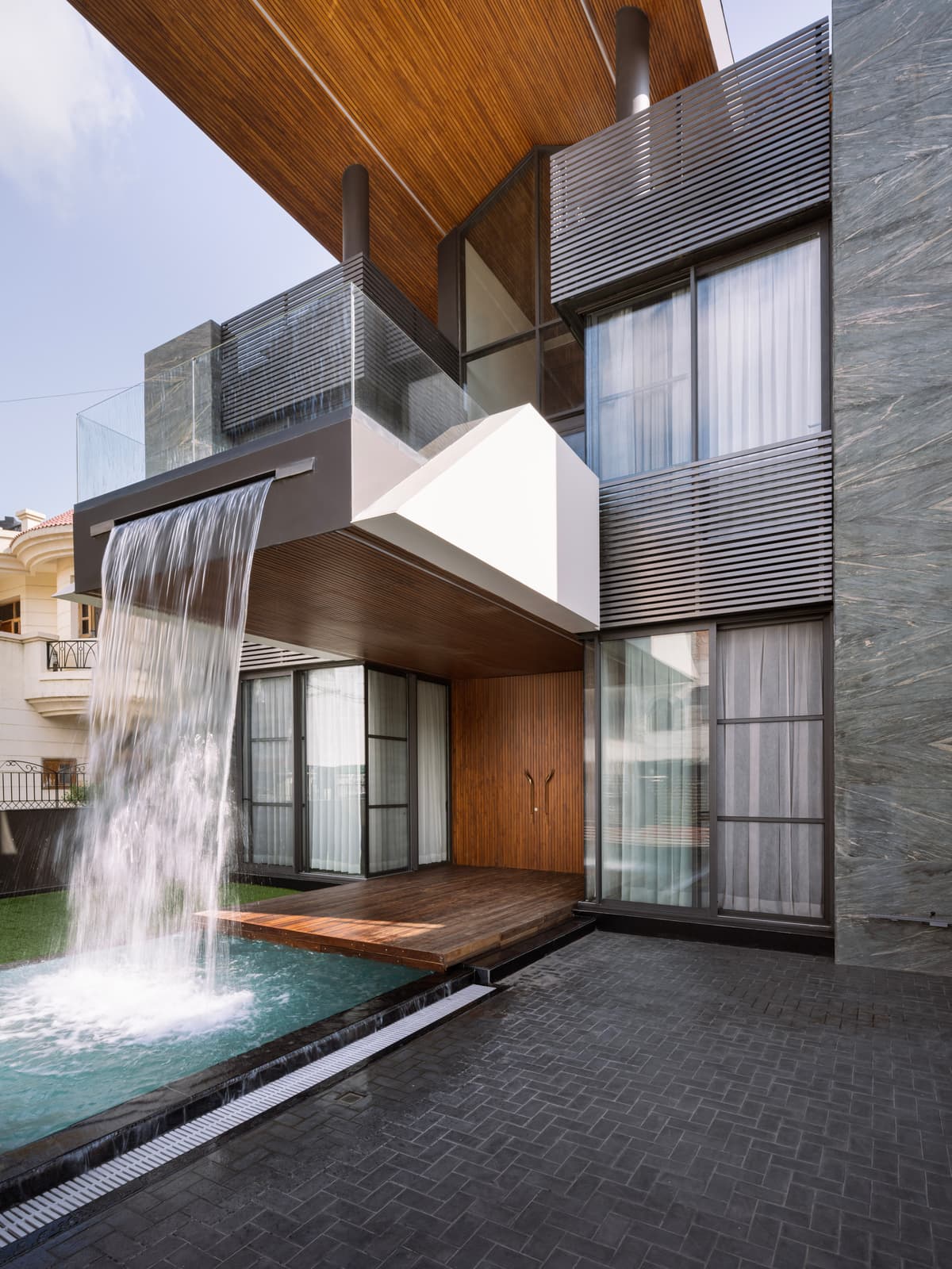 Dramatic cantilevered balcony with cascading water feature, rich wood textures, and vertical fins create a dynamic, inviting entrance experience.