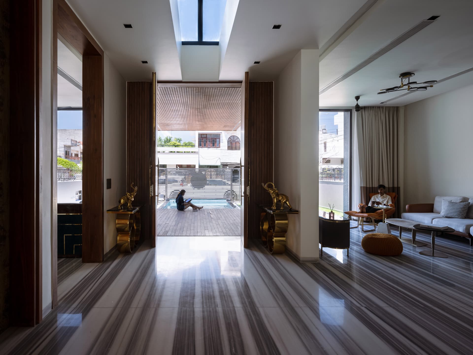 Grand double-height foyer with skylight, fluted wood accents, and striped marble flooring opens seamlessly to a serene outdoor poolscape.