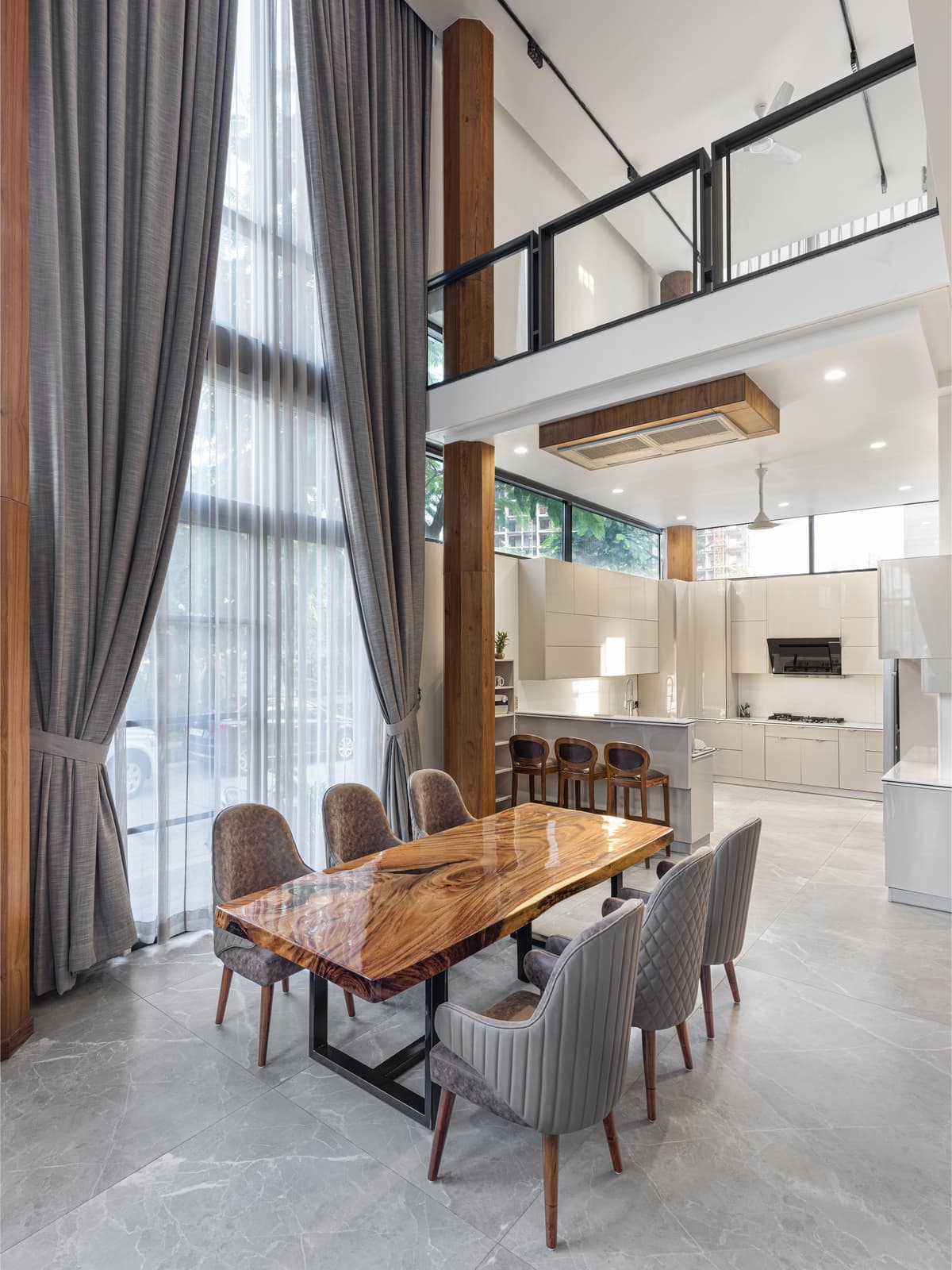 Double-height dining area features dramatic glazing, sheer drapery, a sculptural live-edge wood table, and a sleek modern kitchen beyond.