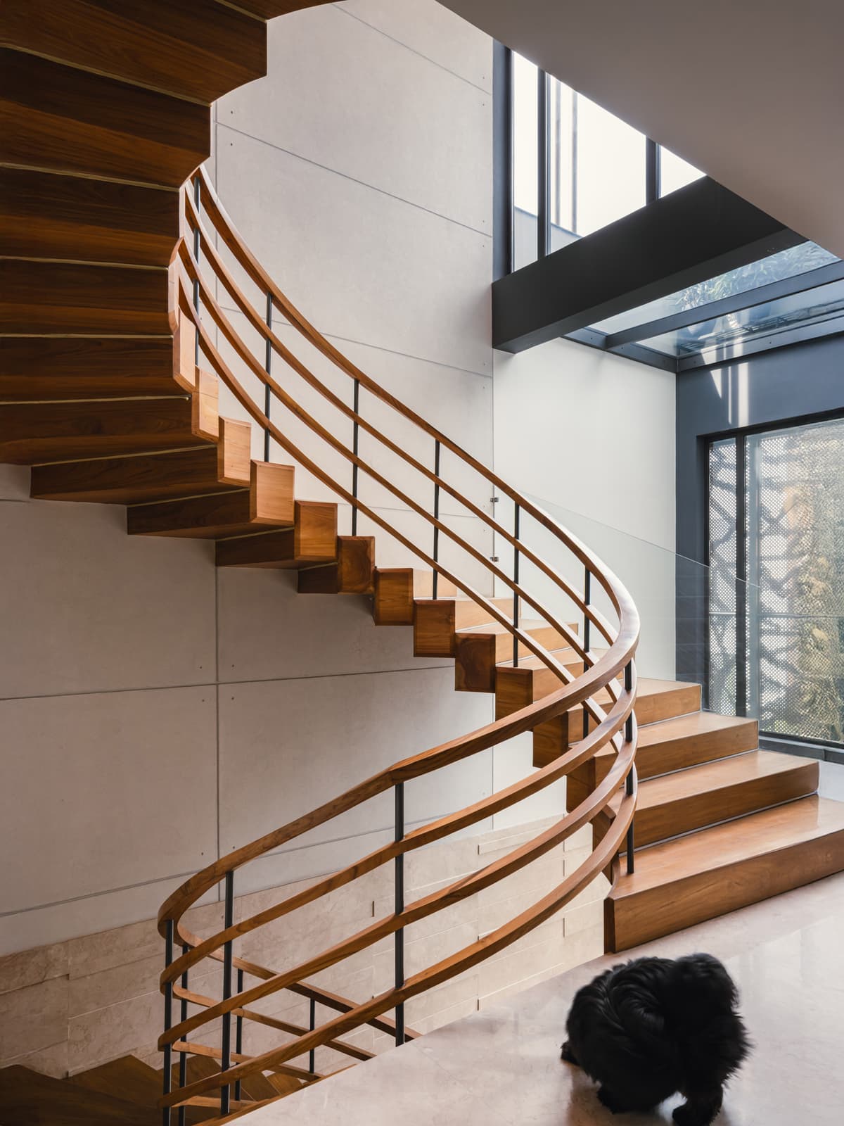 A sculptural wooden staircase with curved railing, open risers, and natural lighting from skylights and large windows enhances vertical movement.
